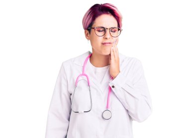 Pembe saçlı, doktor üniforması giyen, diş ağrısı ya da diş hastalığından dolayı ağızlarına acı dolu bir ifadeyle dokunan genç ve güzel bir kadın. dişçi 