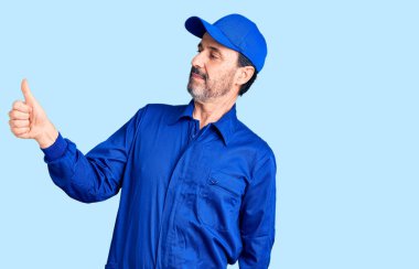 Orta yaşlı, yakışıklı, tamirci üniforması giyen gururlu, gülümseyen, baş parmak kaldıran bir adam. 