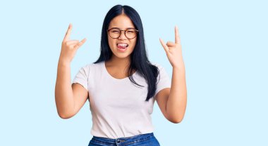 Genç ve güzel Asyalı kız, günlük kıyafetler ve gözlük takmış çılgın bir ifadeyle bağırıyor elleri havada rock sembolü yapıyor. Müzik yıldızı. ağır müzik konsepti. 