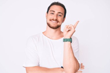 Yüzünde kocaman bir gülümseme olan, el ve parmak ile kameraya bakan, sıradan beyaz tişört giyen genç yakışıklı bir adam.. 