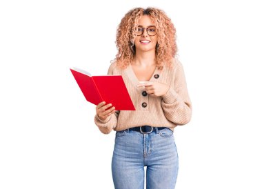 Kıvırcık saçlı, gözlüklü, kitap okuyan, gülümseyen, el ve parmağıyla işaret eden genç sarışın kadın. 