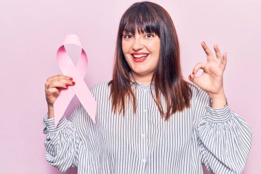 Elinde pembe kanser kurdelesi olan genç ve cüsseli bir kadın parmaklarıyla imza atıyor, gülümseyerek mükemmel bir işaret veriyor. 