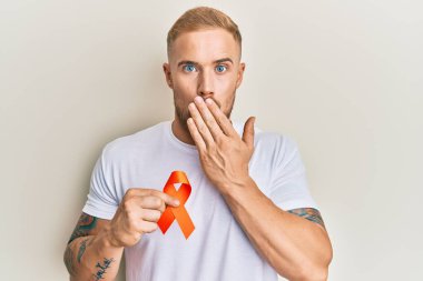 Lösemi için turuncu kurdele tutan genç, beyaz bir adam ağzını eliyle kapatıyor, şok geçiriyor ve hata yapmaktan korkuyor. şaşırmış ifade 