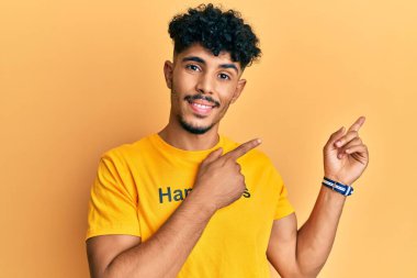Mutluluk mesajı içeren tişört giyen genç Arap yakışıklı adam gülümseyip kameraya bakıyor ve iki eliyle ve parmaklarıyla kenarı işaret ediyor.. 