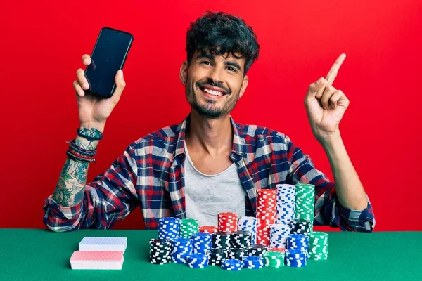 Mladý Hispánec Sedí Stole Pokerovými Žetony Kartami Držícími Smartphone Úsměvem — Stock fotografie