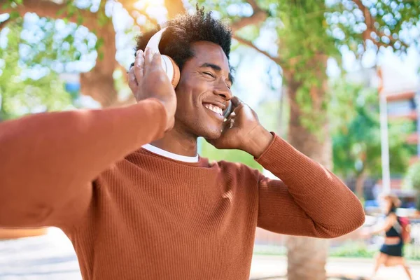 年轻英俊的非洲裔美国男人用耳机听音乐 站在街上 脸上挂着笑容 — 图库照片
