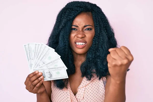 Beautiful african woman holding dollars annoyed and frustrated shouting with anger, yelling crazy with anger and hand raised