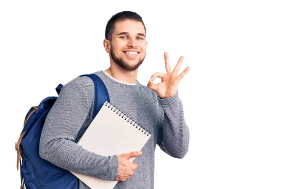 年轻英俊的男子背着学生背包 拿着笔记本 手拿着手签 微笑着表示友善的手势 是极好的象征 — 图库照片