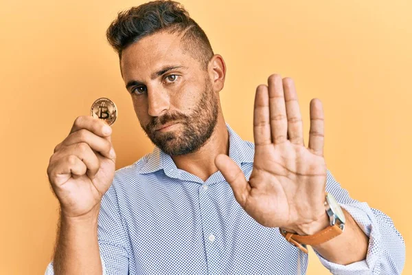 Handsome man with beard holding virtual currency bitcoin with open hand doing stop sign with serious and confident expression, defense gesture