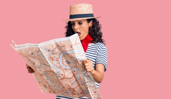 Middle age beautiful woman wearing hat and scarf holding city map scared and amazed with open mouth for surprise, disbelief face
