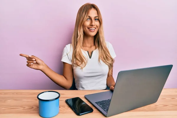 漂亮的金发女人在用电脑笔记本电脑工作时 高兴地微笑着 手指手画脚地指向旁边 — 图库照片