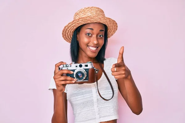 Young african american woman wearing summer hat holding vintage camera pointing fingers to camera with happy and funny face. good energy and vibes.