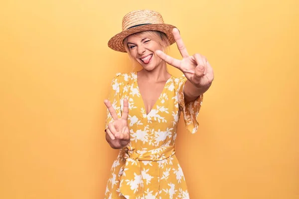 Beautiful blonde woman on vacation wearing summer hat and dress over yellow background smiling with tongue out showing fingers of both hands doing victory sign. Number two.