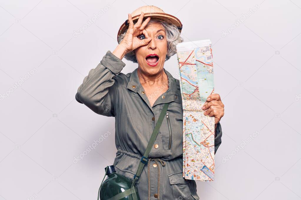 Senior grey-haired woman wearing explorer hat holding map smiling happy doing ok sign with hand on eye looking through fingers 