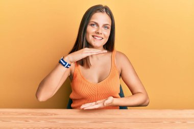 Kafkasyalı genç bir kadın, gündelik giysiler giyip masanın üzerine oturmuş büyük ve büyük beden işareti, ölçü sembolü göstererek el kol hareketi yapıyor. Kameraya bakıp gülümsüyor. ölçüm kavramı. 