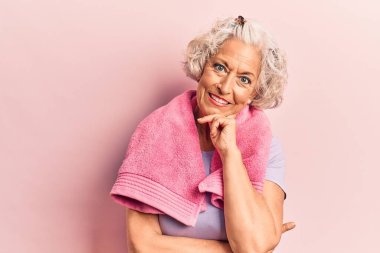 Spor kıyafetleri ve havlu giymiş yaşlı, gri saçlı kadın kameraya kollarını kavuşturup çenesine dokunarak gülümsüyor. Olumlu düşünmek. 