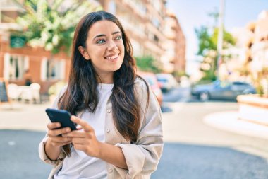 Genç İspanyol kadın şehirde akıllı telefon kullanarak mutlu bir şekilde gülümsüyor..