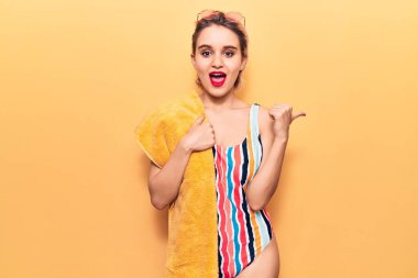 Plaj havlusu tutan, mayo giyen genç, sarışın bir kadın başparmağıyla yan tarafa bakıyor ve ağzı açık gülümsüyor. 