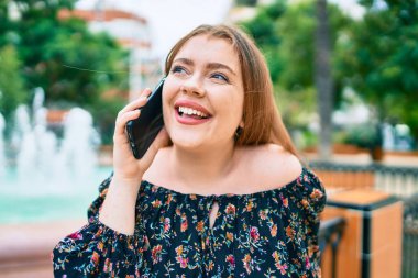 Genç kızıl saçlı kadın parkta akıllı telefondan konuşuyor.