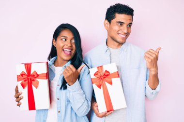 Güzel, genç, Latin bir çift yıldönümü kutlaması için hediyelerini tutarken baş parmağıyla ağzını açıp gülümsüyor. 