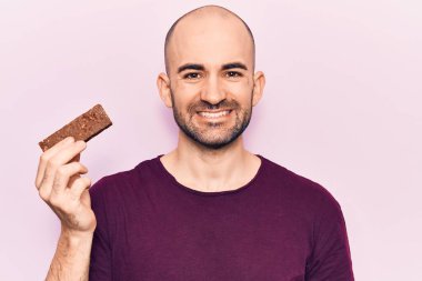 Elinde enerji proteini tutan genç yakışıklı kel adam pozitif ve mutlu görünüyor. Kendine güvenen bir gülümsemeyle ayakta duruyor ve dişlerini gösteriyor. 