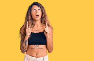 Gündelik giysiler giyen Latin kökenli genç bir kadın şaşırmış ve şaşırmış bir şekilde yukarı baktı ve parmaklarıyla işaret ederek kollarını kaldırdı.. 