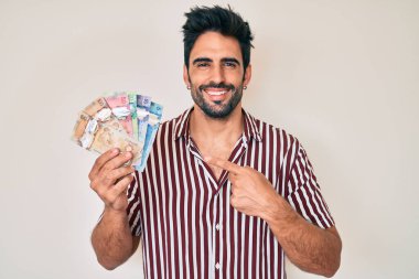 Yakışıklı, sakallı, Kanada doları tutan, eli ve parmağı ile mutlu mesut gülümseyen bir İspanyol. 