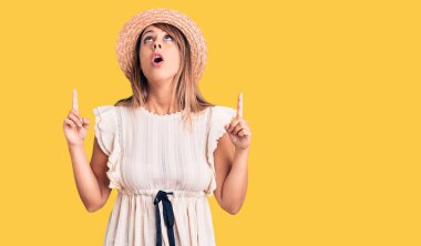 Yaz şapkası ve tişörtü giyen genç ve güzel bir kadın şaşırmış ve şaşırmış bir şekilde yukarı bakıyor ve parmaklarıyla işaret ediyor ve kollarını kaldırıyordu.. 