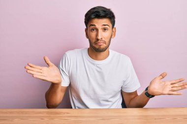 Masada oturan, gündelik beyaz tişört giyen, elleri ve kolları havada, kafası karışmış genç, yakışıklı bir adam. şüphe kavramı. 