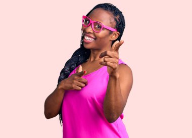 Sıradan kıyafetler giyen ve gözlüklü genç Afro-Amerikan kadın mutlu ve komik yüzlü parmaklarını kameraya doğrultuyor. iyi enerji ve titreşimler. 