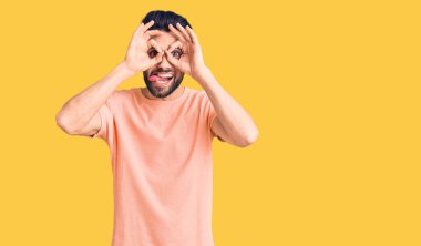 Sakallı, sıradan tişörtlü genç yakışıklı bir adam dürbün gibi jest yapıyor, dili dışarı çıkarıyor, gözleri parmaklarından bakıyor. çılgın ifade. 