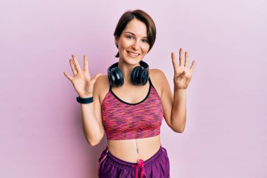 Kısa saçlı, spor giyimli, kulaklık takan genç esmer kadın dokuz numaralı parmaklarıyla kendini beğenmiş ve mutlu gülümserken kendini gösteriyor ve işaret ediyor.. 