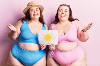 Güneş tutan bikini giyen genç ve büyük beden ikizler zaferi mutlu bir gülümsemeyle kutluyor ve kazanan el kaldırarak ifade ediyor. 