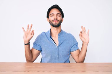 Masaya oturmuş, rahat rahat gülümseyen, gözleri kapalı meditasyon yapan ve parmaklarıyla meditasyon yapan yakışıklı İspanyol adam. yoga konsepti. 