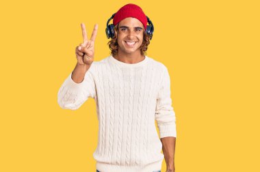 Genç İspanyol adam kulaklık kullanarak müzik dinliyor. İki numaralı parmaklarıyla işaret ediyor. Kendinden emin ve mutlu bir şekilde gülümsüyor.. 