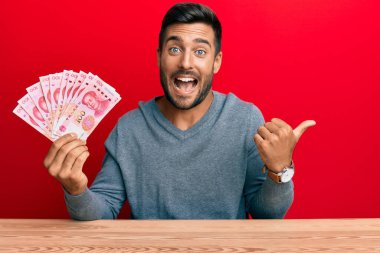 Yakışıklı İspanyol adam Yuan Çin banknotlarını tutuyor başparmağıyla yan tarafı işaret ediyor ve ağzı açık gülümsüyor. 