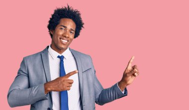 Yakışıklı Afro saçlı, iş ceketi giyen, gülümseyen ve kameraya bakan, iki eli ve parmakları yanda olan Afro Amerikalı bir adam.. 