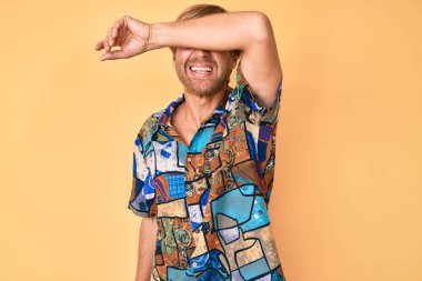 Yazlık tişört giyen, kollarıyla gözlerini kapatan neşeli ve komik gülümseyen genç sarışın adam. kör kavram. 