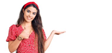 Yazlık elbise giyen esmer kız avuç içi eli gösteriyor ve baş parmaklarıyla iyi bir jest yapıyor, mutlu ve neşeli gülümsüyor. 