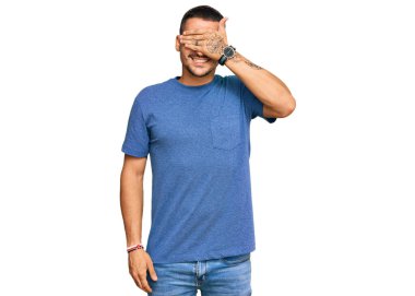 Dövmeleri olan, sıradan kıyafetler giyen, gülümseyen ve elleri yüzünde, şaşırtmak için gözlerini kapatan yakışıklı bir adam. kör kavram. 