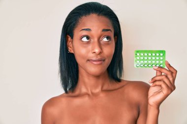 Genç Afrikalı Amerikalı kadın elinde doğum kontrol hapları tutarken gülümsüyor ve diğer tarafa bakıyor.. 