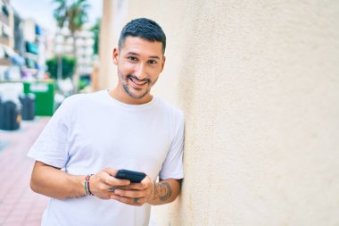 Duvara dayanmış akıllı telefon kullanan, mutlu gülümseyen genç İspanyol adam..