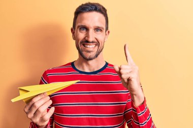 Sarı kâğıt uçak tutan genç yakışıklı bir adam izole arka planda duruyor ve bir fikir veya soru işareti ile gülümsüyor mutlu bir yüzle, bir numara