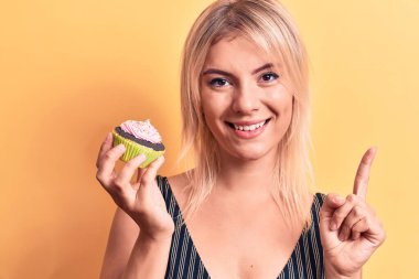 Sarışın, güzel bir kadın izole edilmiş sarı arka planda tatlı çikolatalı kek yiyor. Bir fikir ya da soru işareti ile gülümsüyor.