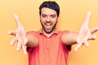 Gündelik giysiler giymiş genç İspanyol adam kameraya bakıp sarılmak için kollarını açmış gülümsüyor. Mutluluğu kucaklayan neşeli bir ifade.. 