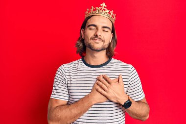 Prens tacı takan genç yakışıklı adam elleri göğsünde gülümsüyor, gözleri kapalı ve yüzünde minnet ifadesi var. sağlık kavramı. 