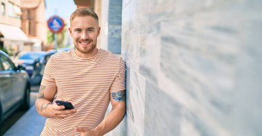 Genç İrlandalı mutlu bir şekilde gülümsüyor. Şehrin duvarına yaslanmış akıllı telefon kullanıyor..