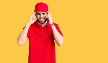 Sakallı, teslimat üniforması giyen, eli başının üstünde, başı ağrıyan genç, yakışıklı bir adam. migren ağrısı. 