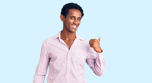 アフリカのハンサムな男身に着けていますカジュアルなピンクのシャツ笑顔で幸せな顔を見て 親指アップで側面を指して — ストック写真