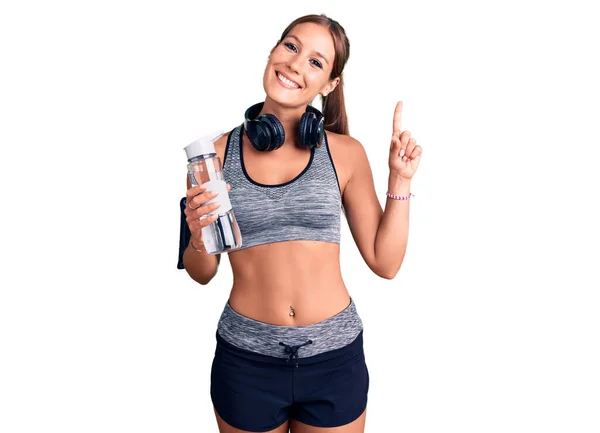 若い美しいヒスパニック女性を身に着けているスポーツウェアを保持水ボトル驚きでアイデアや質問を指差す指で幸せな顔 ナンバーワン — ストック写真